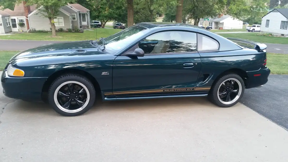 A dark green 1990s Ford Mustang GT is parked on a residential driveway, showing its side profile with gold racing stripes…