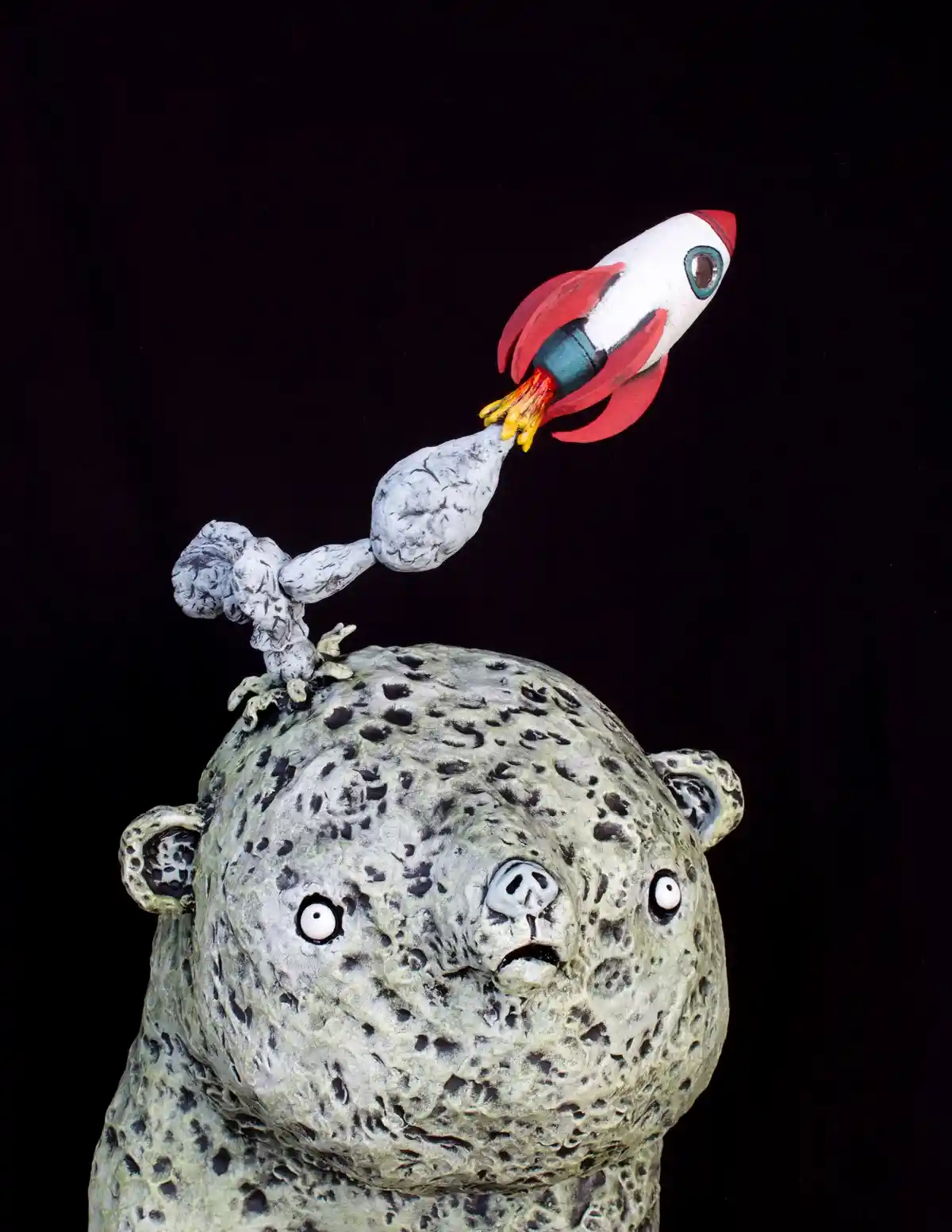 A textured, gray, bear-like sculpture with wide, startled eyes and a surprised expression has a small, cartoonish…