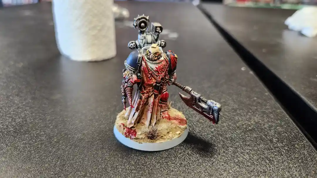 A heavily painted miniature figurine of a warrior in ornate armor, covered in red paint resembling blood, stands on a…