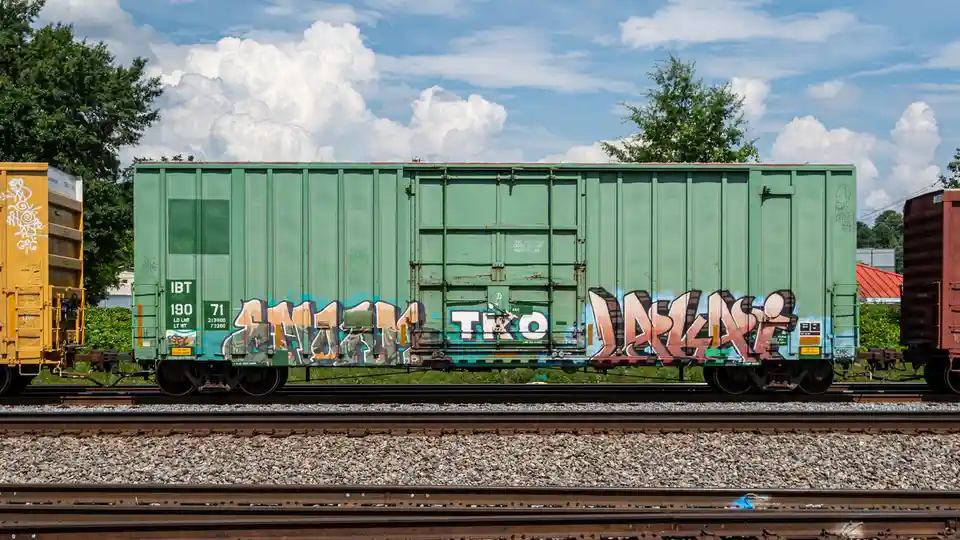 A green freight train car covered in graffiti sits on tracks under a cloudy sky, with text overlaid reading 'this car excess…