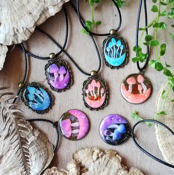 Several handmade necklaces with oval or round pendants featuring colorful mushroom illustrations in resin, displayed on a…