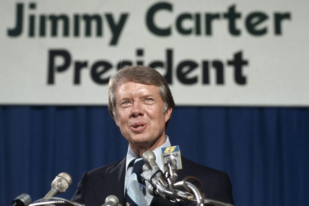 A man in a suit and tie stands at a podium with multiple microphones, speaking in front of a backdrop that reads 'Jimmy…