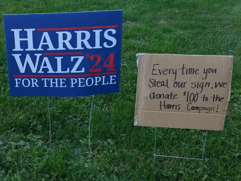 Two campaign signs stand in grass: a blue 'HARRIS WALZ '24 FOR THE PEOPLE' sign and a cardboard sign with handwritten text…