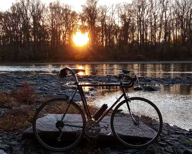 A black road bicycle with a water bottle is parked on a large rock beside a river at sunset. The sun is low in the sky…