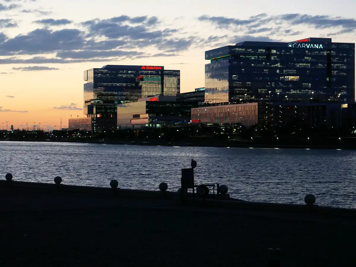 A city skyline at dusk with modern glass buildings along a waterfront, featuring illuminated 'StateFarm' and 'CARVANA' logos…