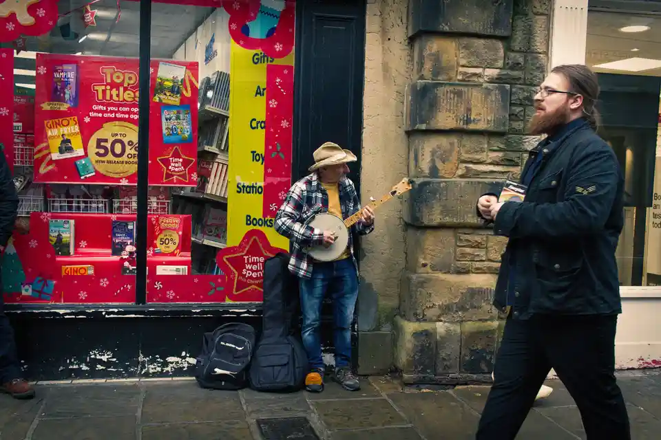 A man with a beard and glasses walks past a street musician playing a banjo outside a bookstore decorated with…
