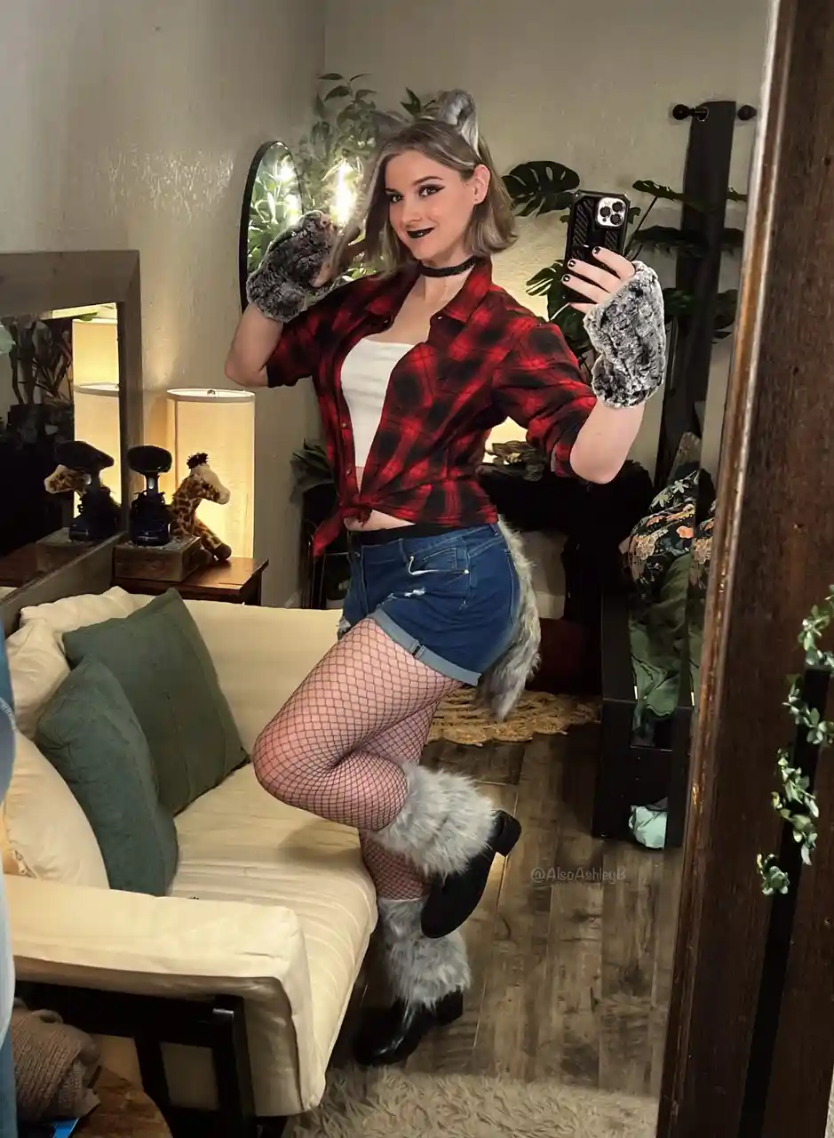 A woman with grey hair and wolf ears takes a mirror selfie in a living room, wearing a red plaid shirt, denim shorts…