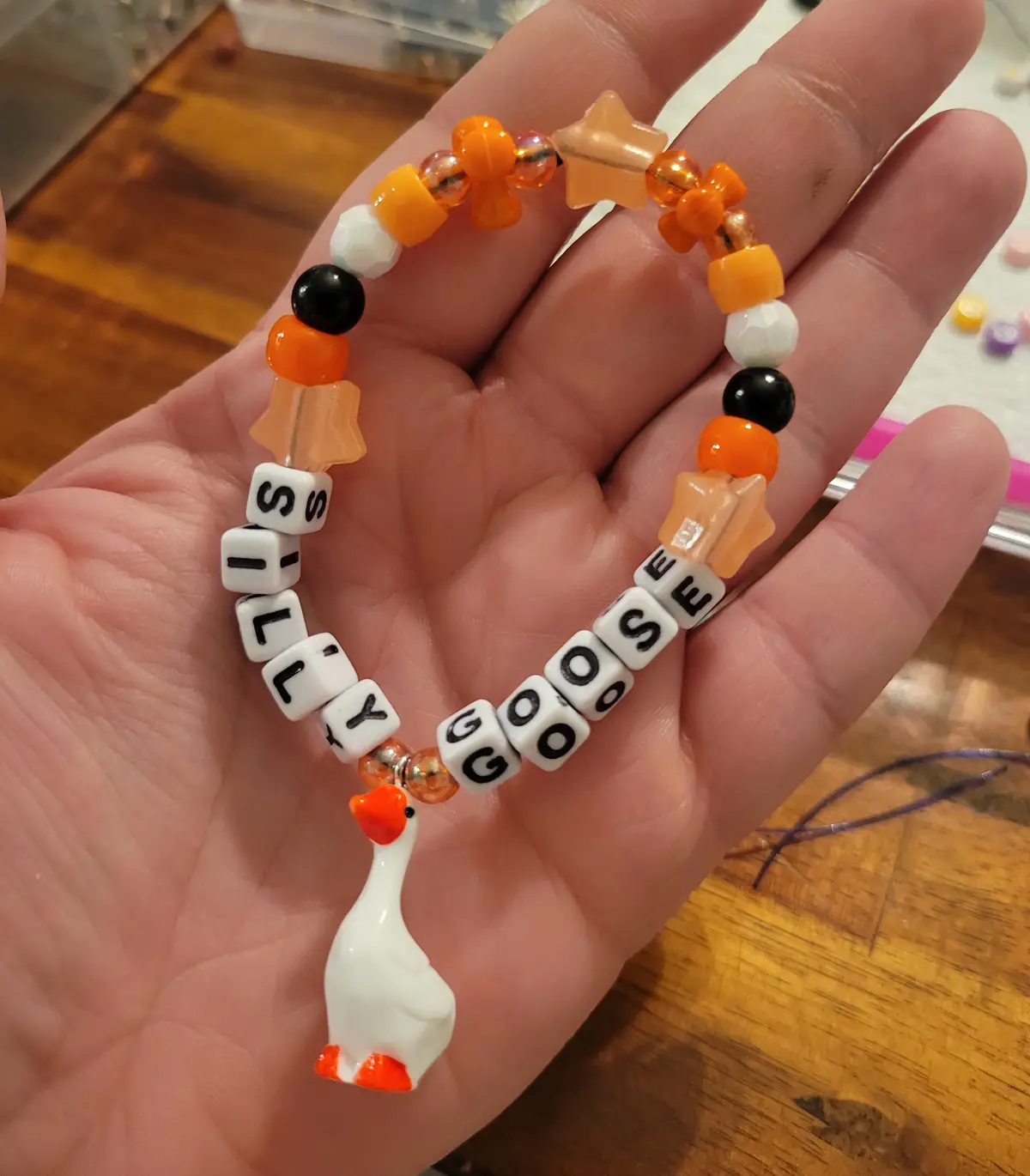 A hand holds a handmade beaded bracelet with orange, black, and white beads, including letter beads spelling 'silly goose'…