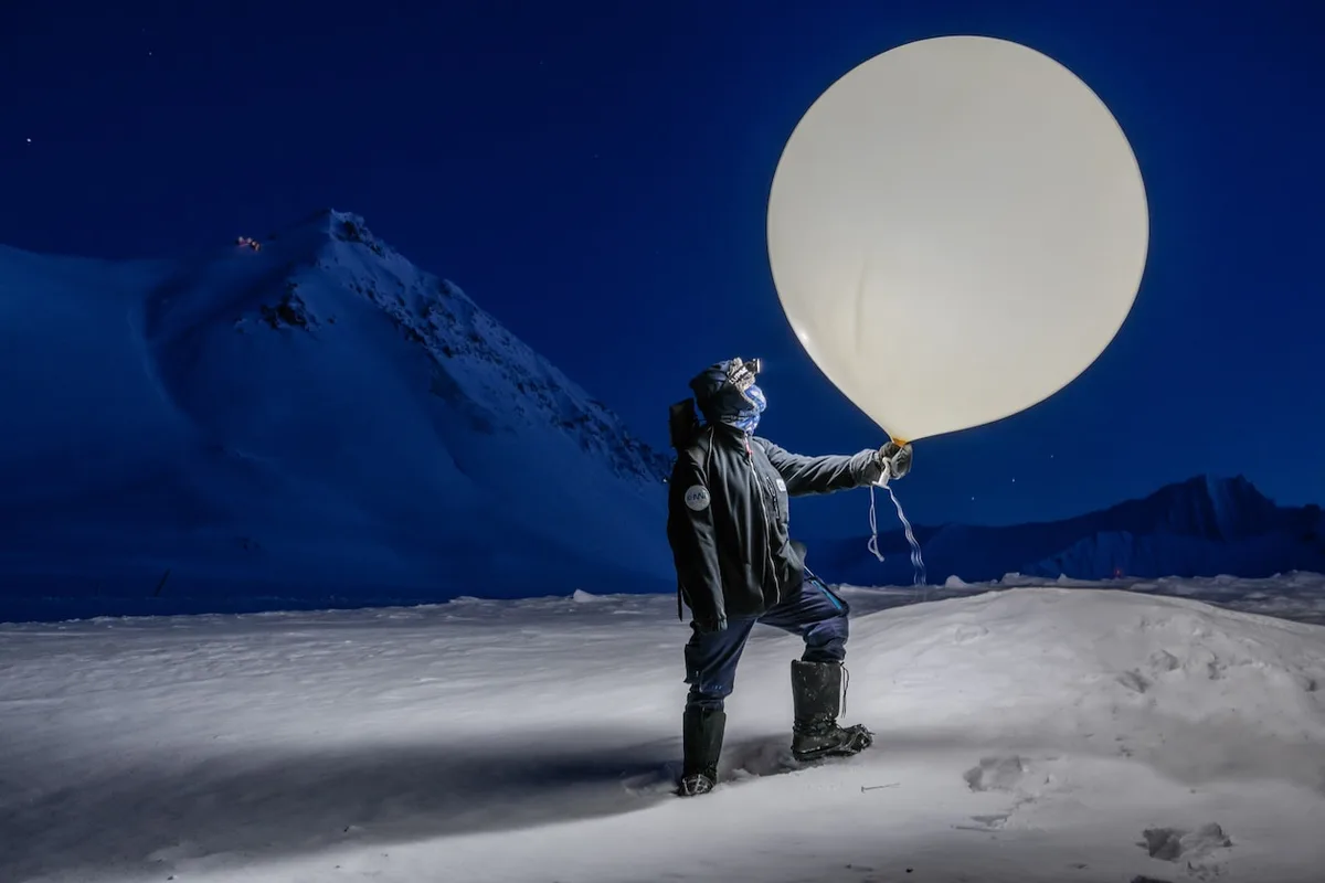 A person in winter gear stands in a snowy, mountainous landscape at night, holding a large white balloon. The sky is dark…