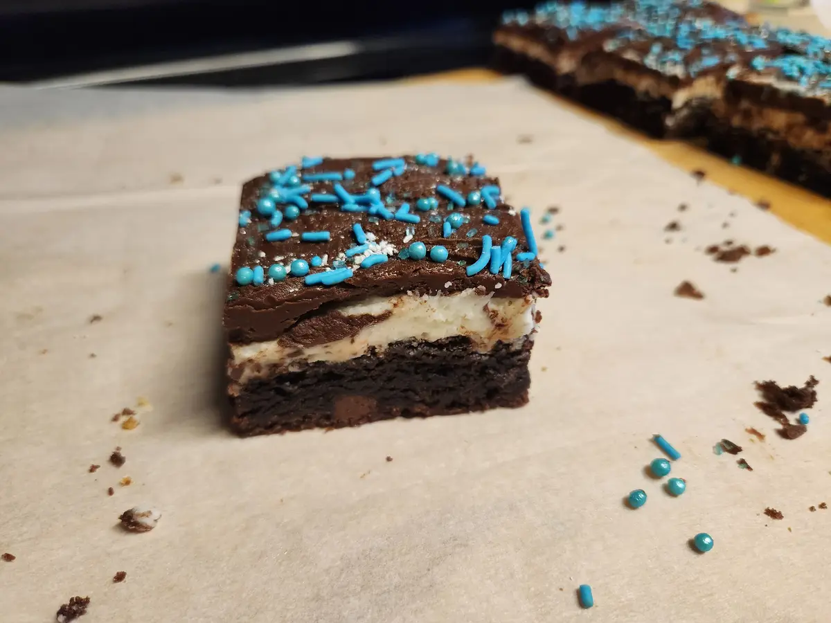 A close-up photo shows a square chocolate brownie with a white cream layer and chocolate frosting, topped with blue…