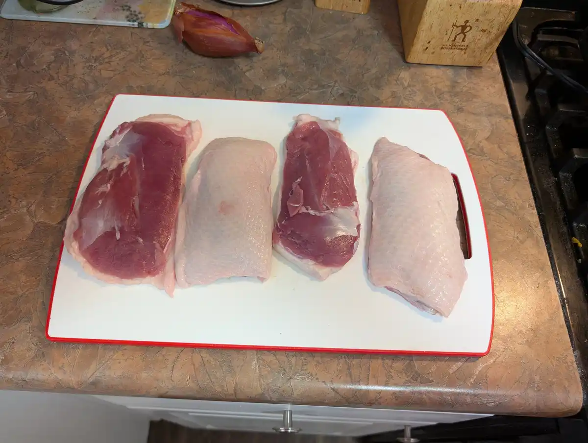 Four raw duck breasts are arranged on a white cutting board with a red border, placed on a brown marbled kitchen countertop…