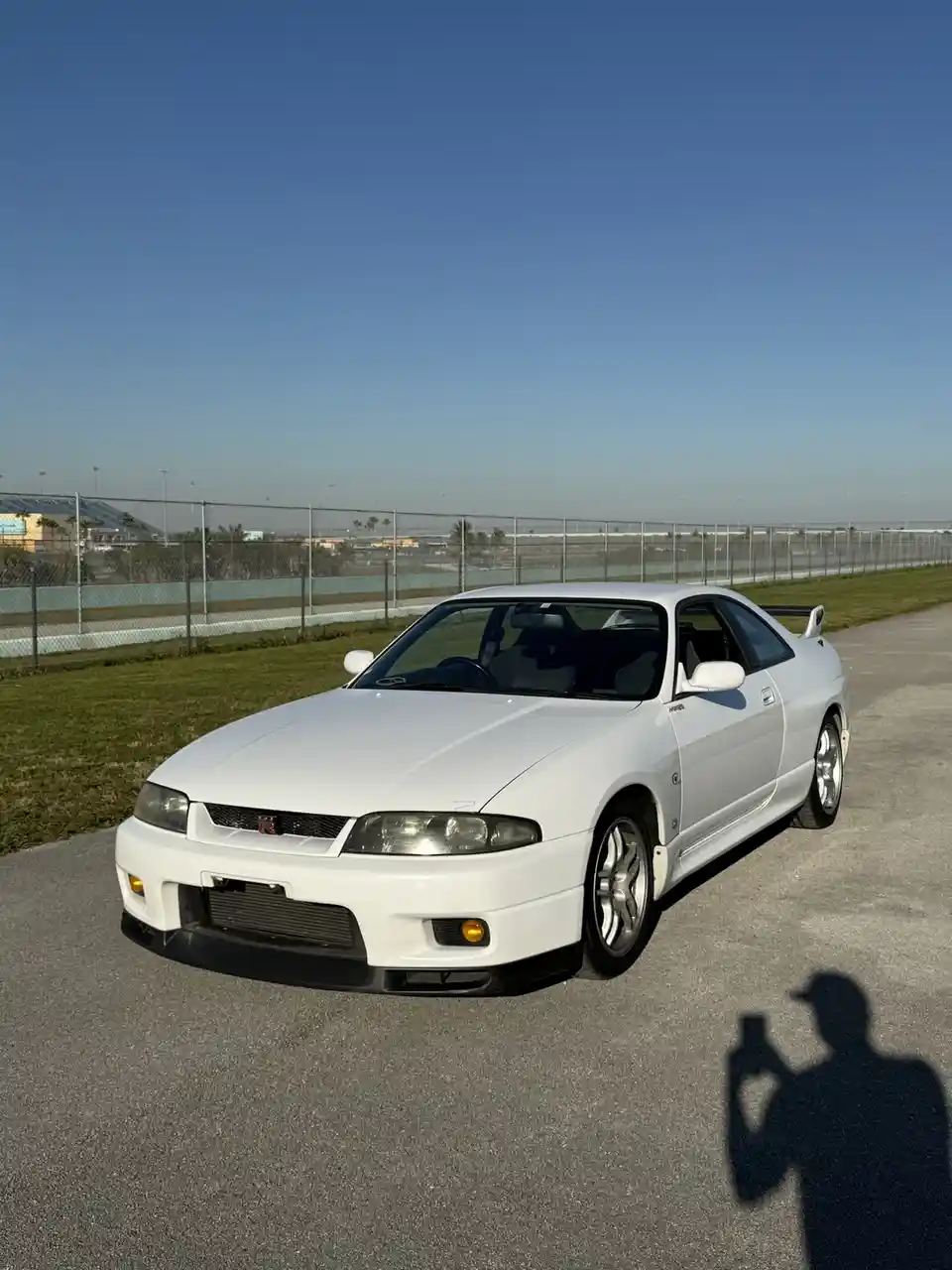 A white Nissan Skyline R34 is parked on an asphalt surface near a chain-link fence with a racetrack in the background under…