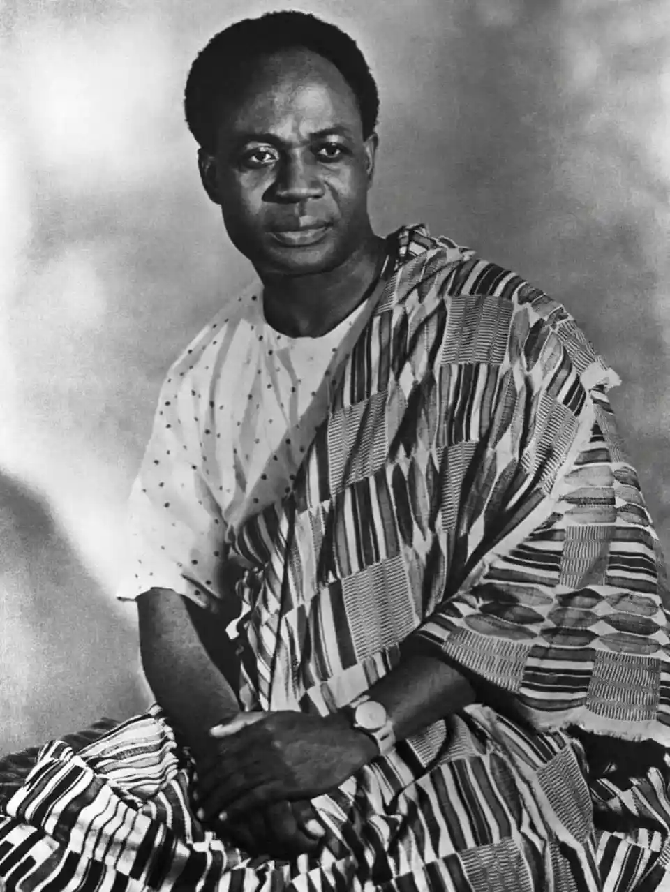 A black and white portrait of a man wearing a patterned garment, looking directly at the camera with a neutral expression…