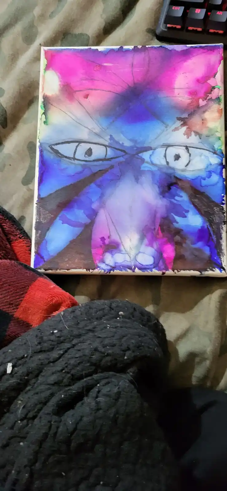 A small, colorful watercolor painting on canvas depicts a stylized face with large, intense eyes and abstract, swirling…