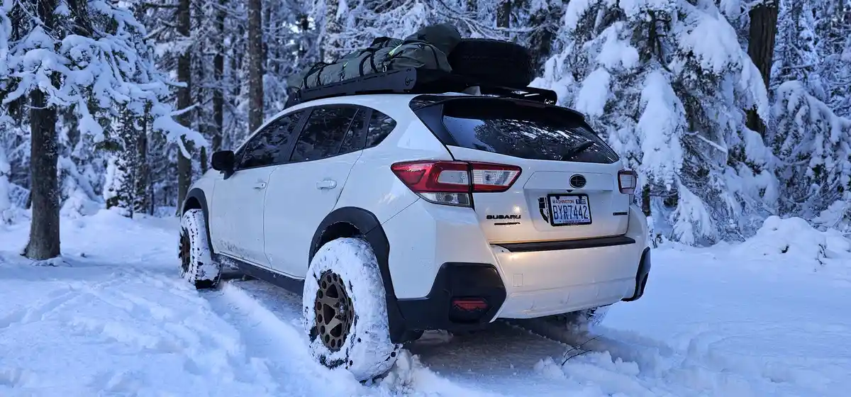 A white Subaru SUV is parked in a snowy forest, its rear and side visible. The vehicle has a roof rack loaded with gear and…