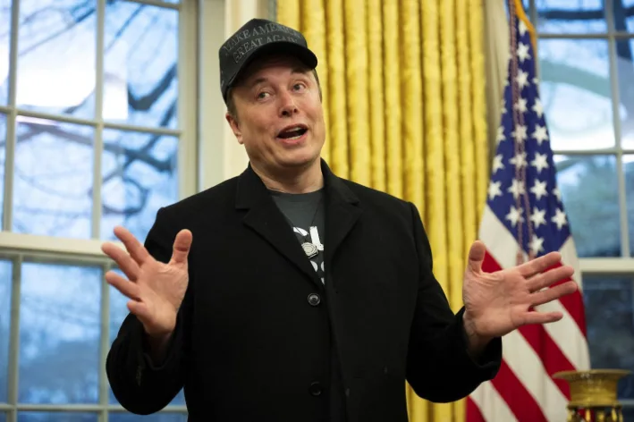 A man wearing a 'MAKE AMERICA GREAT AGAIN' cap and a black coat stands in front of yellow drapes and an American flag…