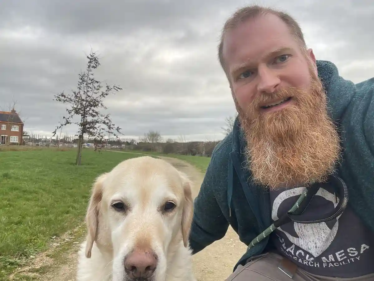 A man with a long red beard wearing a 'Black Mesa Research Facility' t-shirt takes a selfie with a yellow Labrador Retriever…