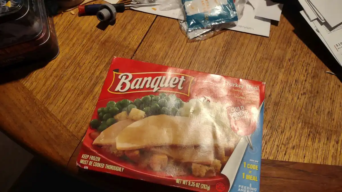 A close-up photo of a Banquet frozen turkey meal box on a wooden table, with a large, translucent plastic knife blade…