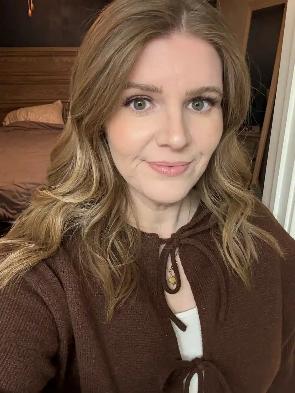 A woman with wavy blonde hair and green eyes smiles at the camera while wearing a brown knitted cardigan over a white top…