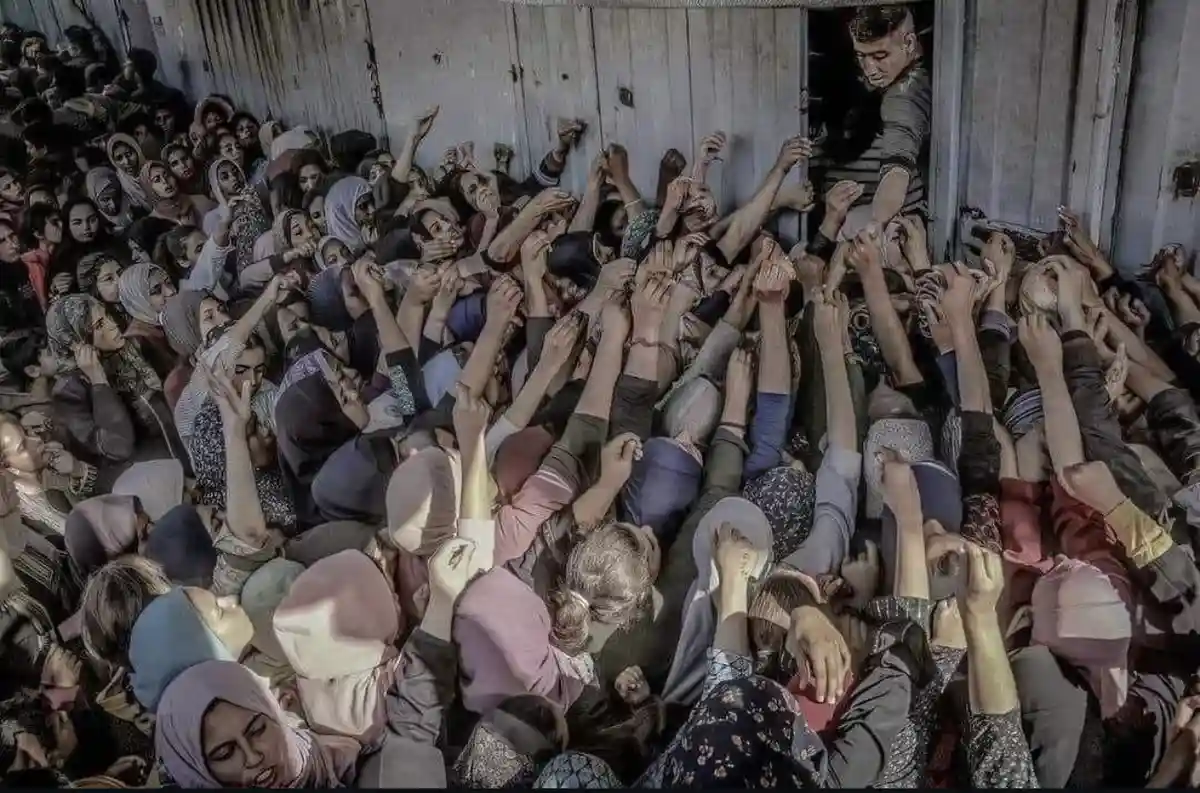 A crowded room filled with people, mostly women wearing headscarves, all reaching desperately toward a single man standing…