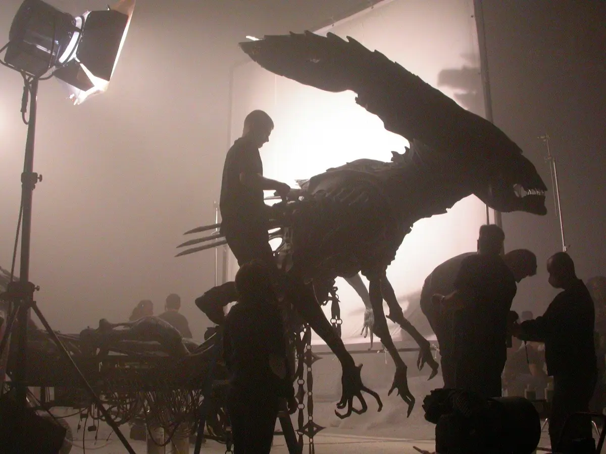 A behind-the-scenes photo shows a large, menacing alien creature prop being assembled by crew members in a dimly lit studio…
