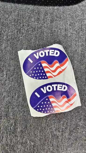 Two identical 'I VOTED' stickers featuring a stylized American flag design are stuck side-by-side on a gray fabric surface…