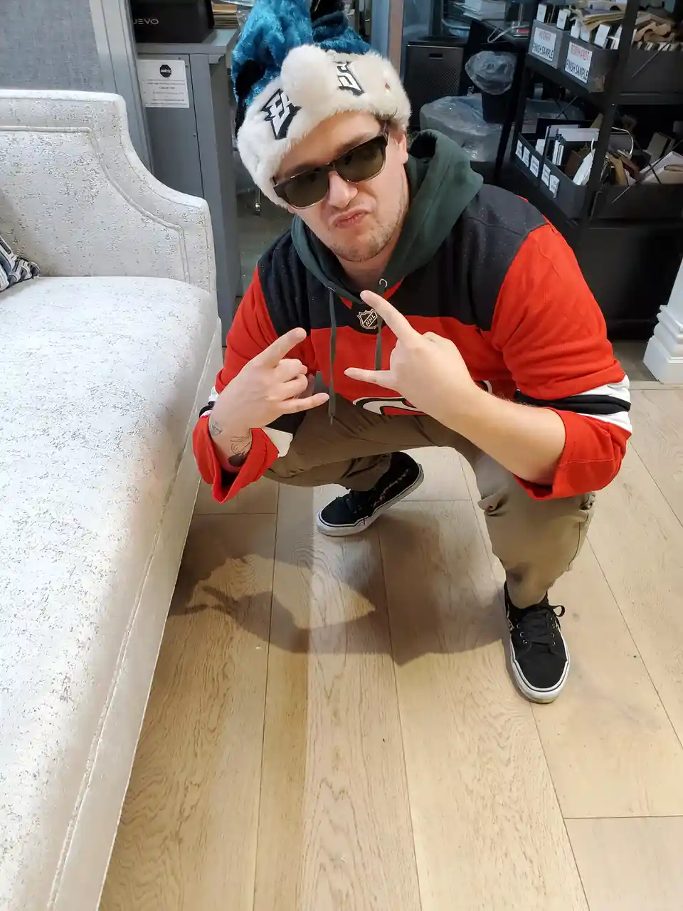 A man wearing a blue and white furry hat, sunglasses, and a red and black hoodie is crouching next to a light-colored couch…