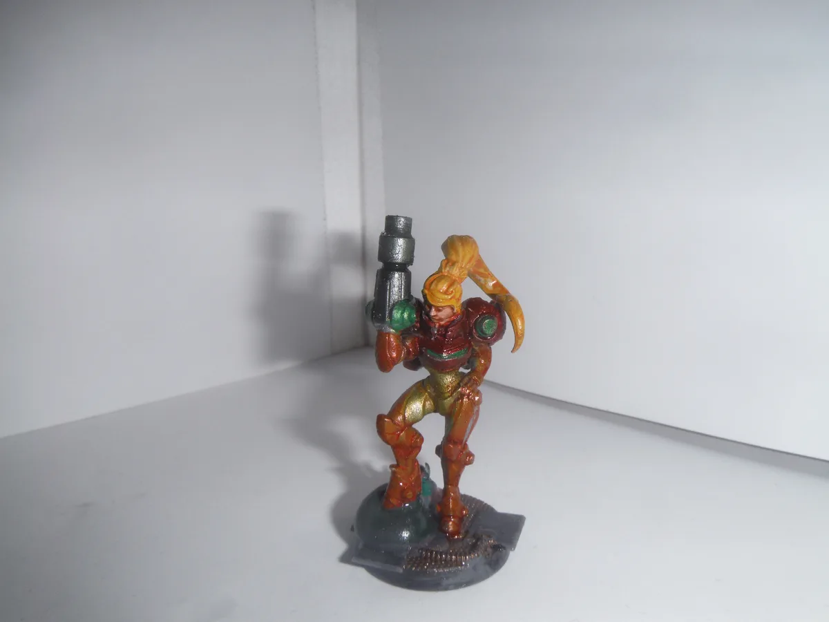A painted figurine of a female video game character in red and gold armor, holding a large gun, stands on a grey base…