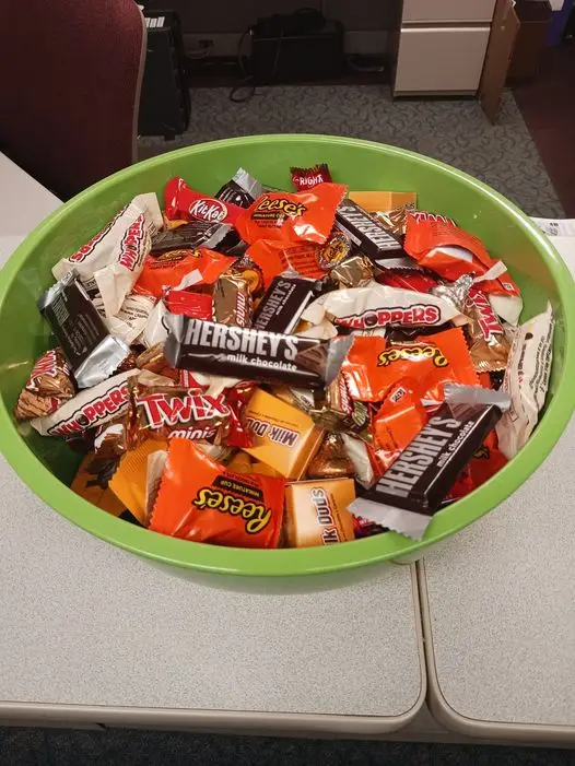 A green plastic bowl overflowing with various individually wrapped chocolate candies including Hershey's, Reese's, Twix, and…