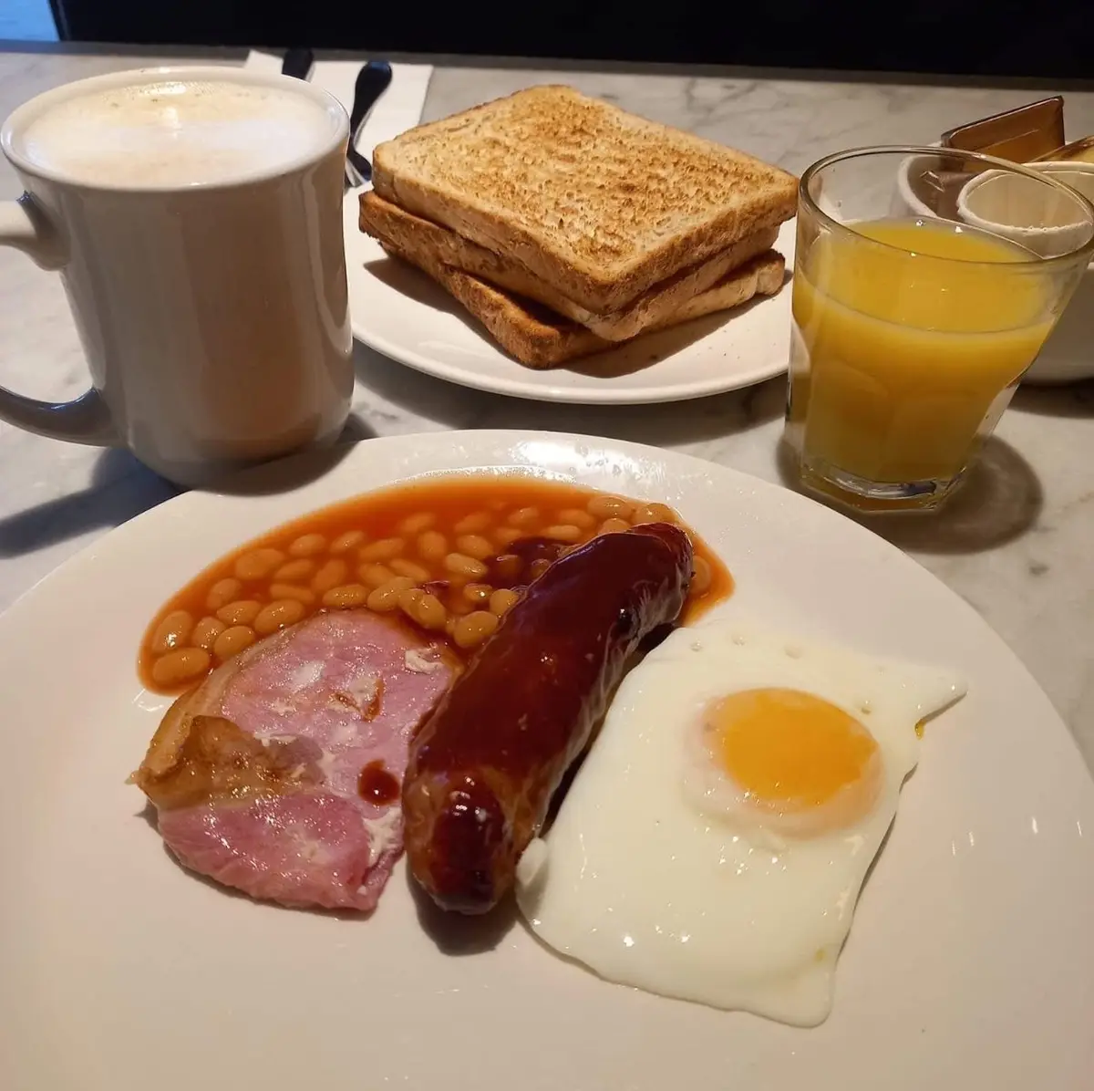 A full English breakfast is laid out on a table, featuring a plate with fried egg, sausage, ham, and baked beans, alongside…