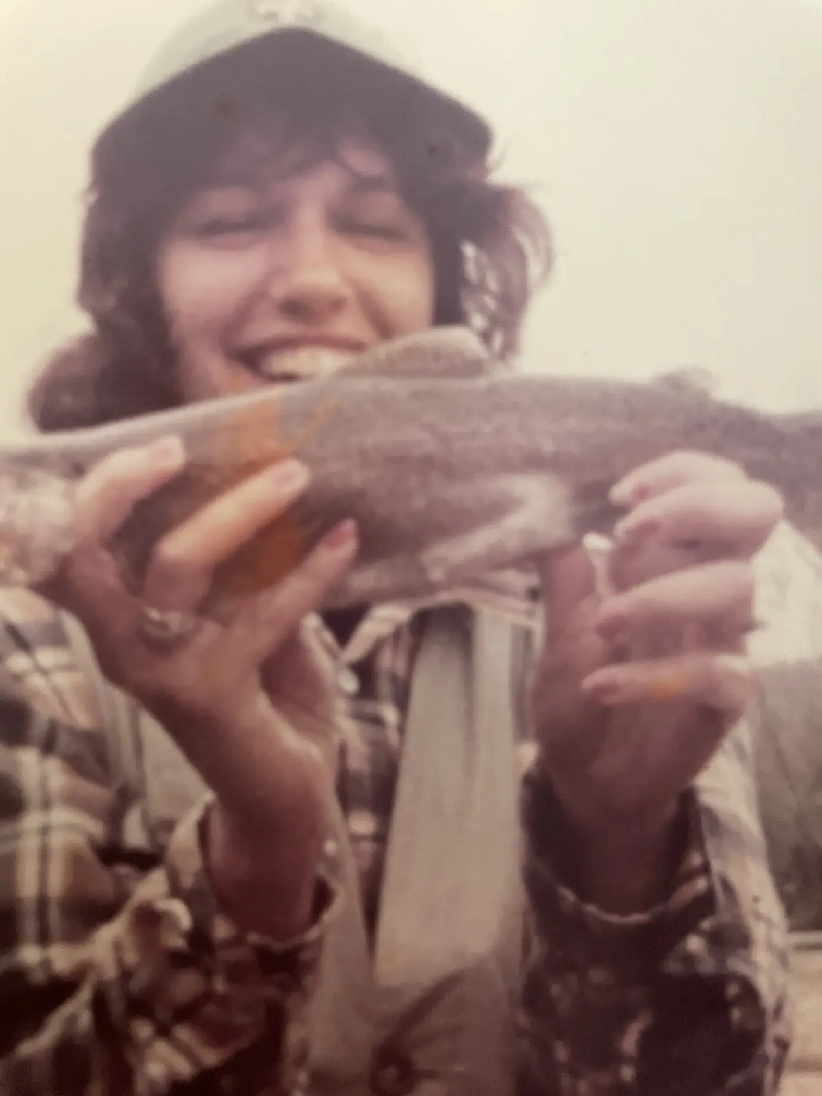 A person with curly hair and a cap, wearing a plaid shirt, is holding up a large fish with both hands, grinning widely with…