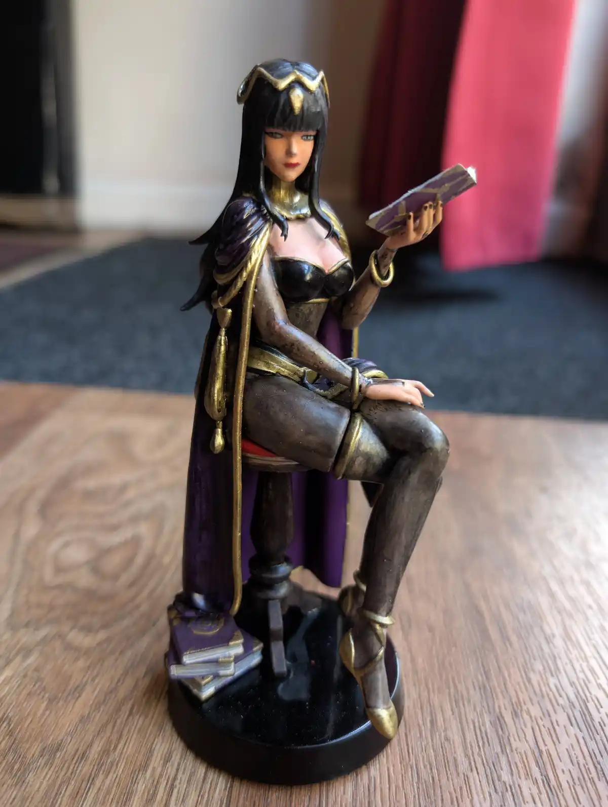 A figurine of a woman with long black hair and gold accessories, wearing a purple and gold outfit, sits on a small stool…