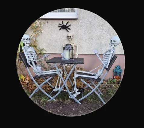 Three plastic skeleton decorations are seated at a small outdoor table with chairs, appearing to be having a conversation…