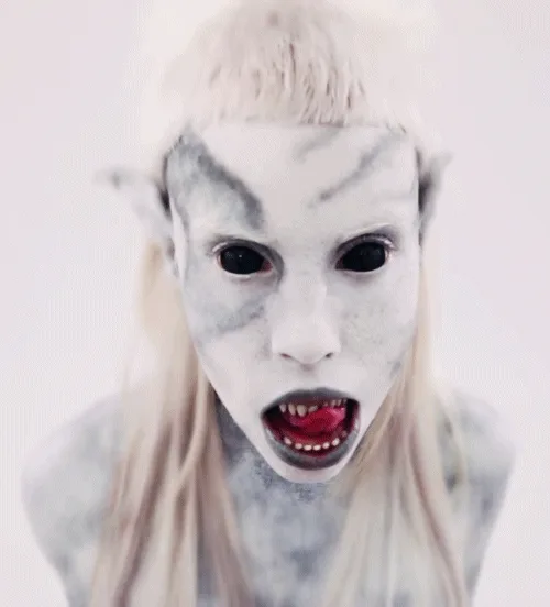 A person with pale, grayish-white face paint, pointed ears, and blonde hair stares intensely with wide eyes and an open…