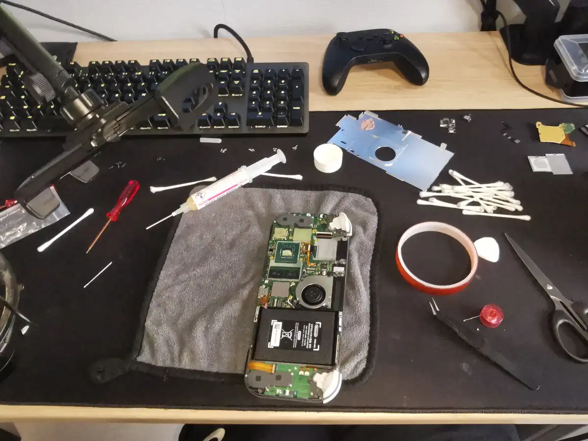 An overhead shot of a cluttered desk with a disassembled electronic device, likely a gaming console, laid open on a grey…