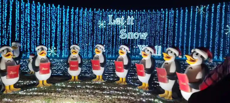 A row of cartoon penguin statues wearing Santa hats and holding red songbooks labeled 'Peace' stand in front of a blue LED…