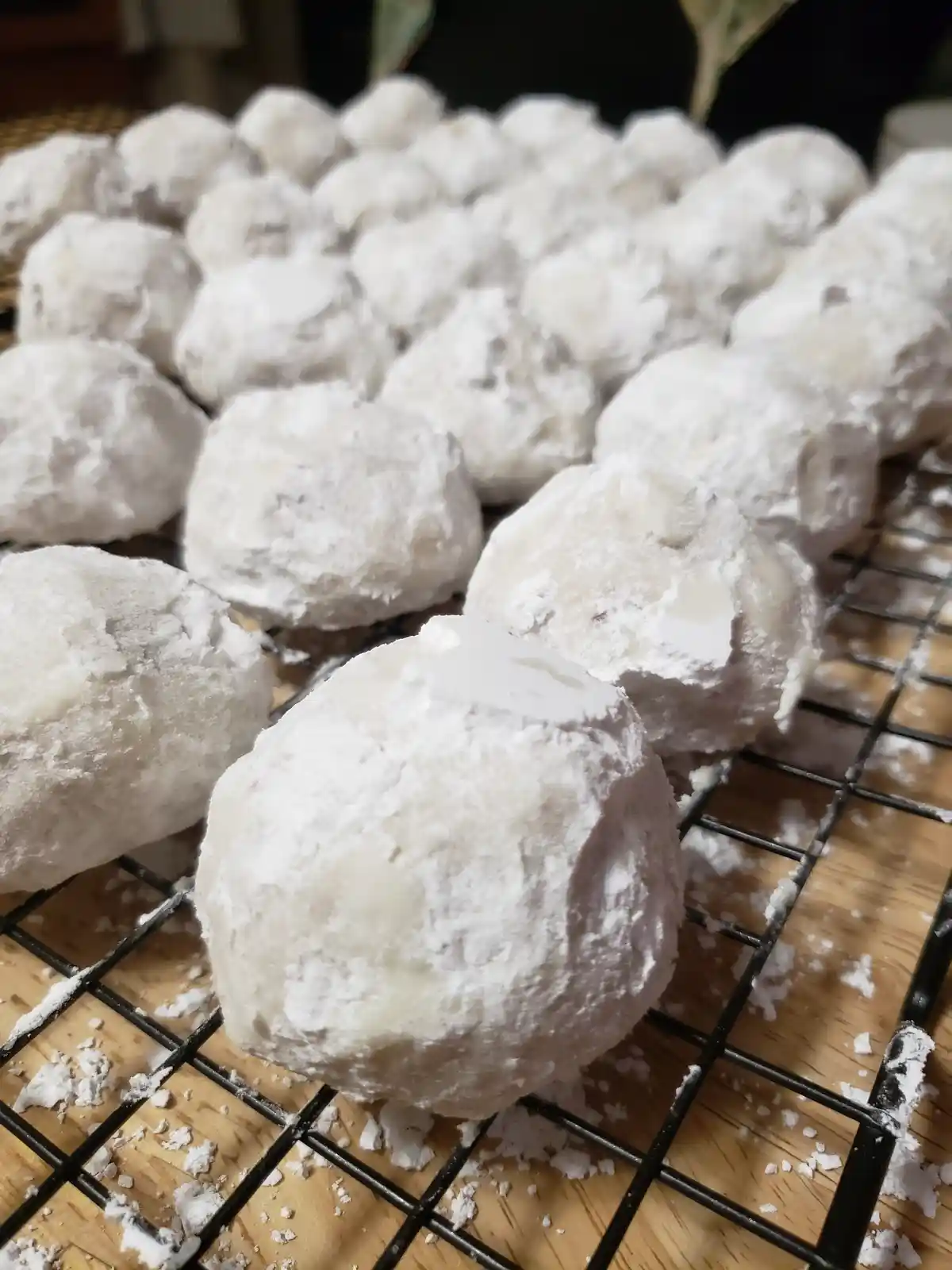 A close-up photo shows many round, white, powdered sugar-coated cookies or confections arranged on a black wire cooling rack…