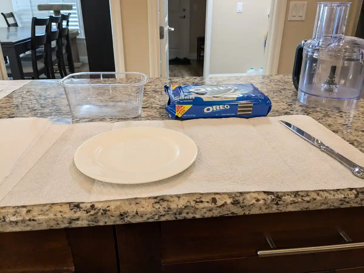 A kitchen counter displays a white plate, a bag of Oreo cookies, a clear plastic container, and a butter knife, all arranged…