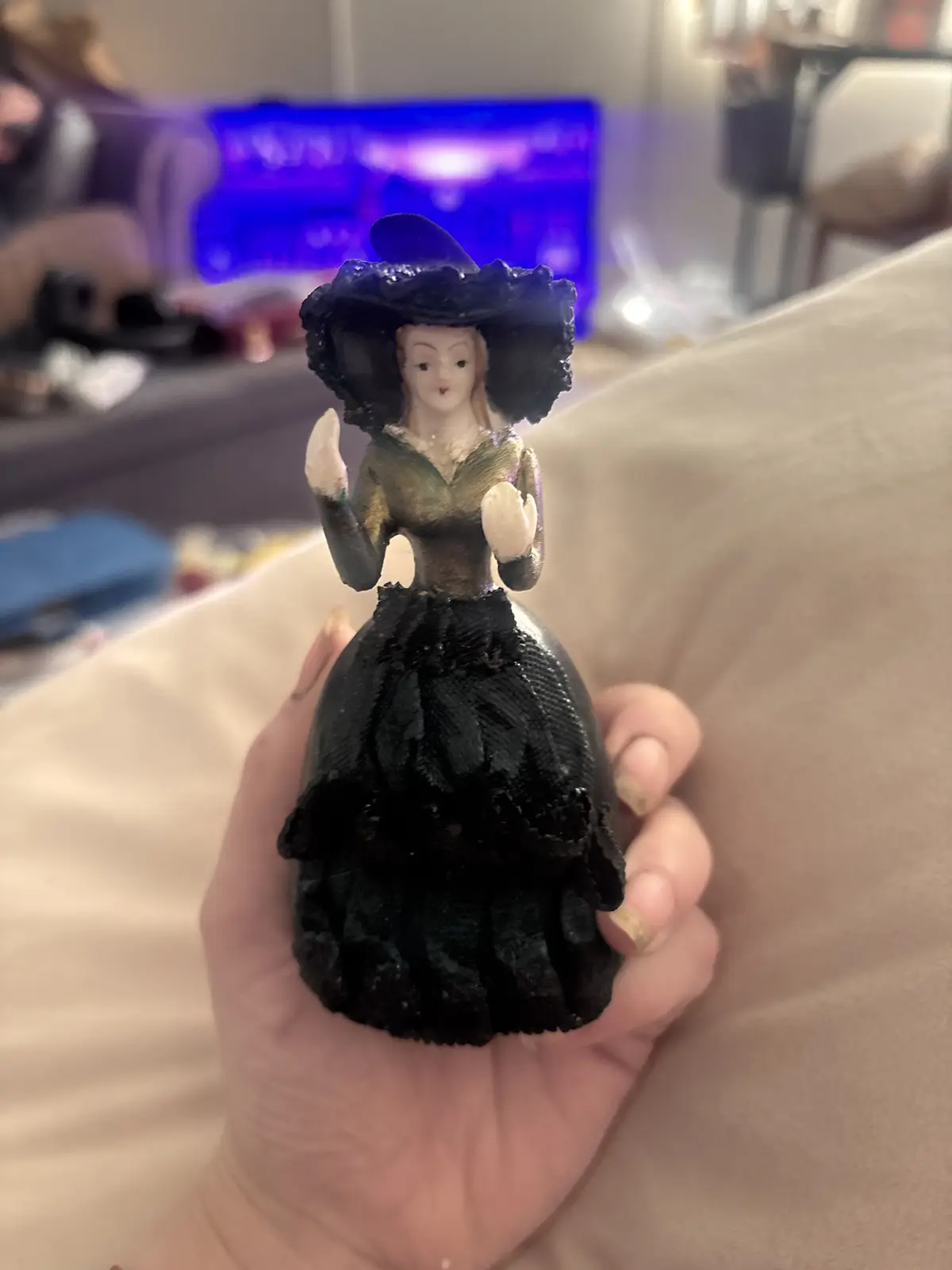 A hand holds a small figurine of a woman dressed in a black Victorian-style gown and hat, with white gloves, against a…