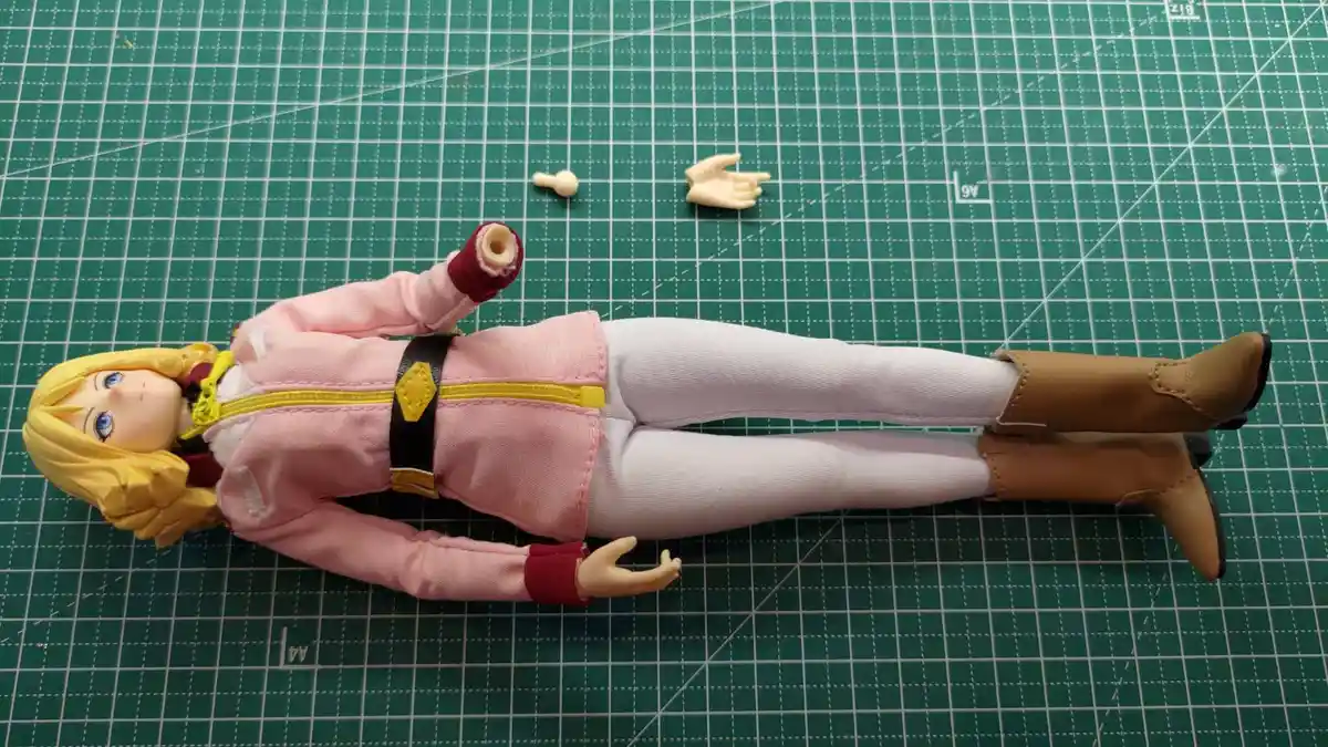 An anime-style action figure with blonde hair and blue eyes lies flat on a green cutting mat, dressed in a pink jacket…