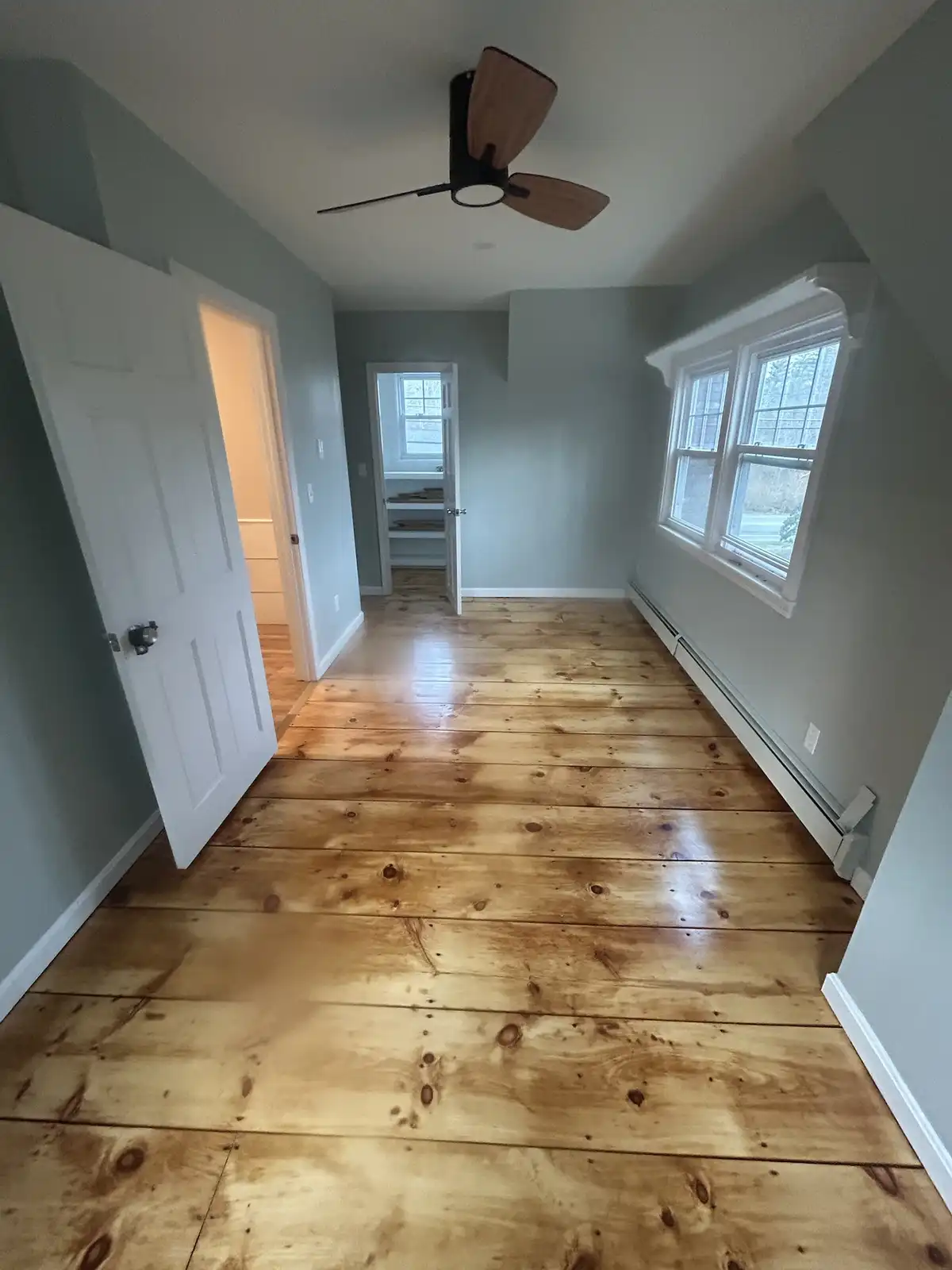 An empty, newly renovated room with light gray walls, white trim, and shiny light-colored hardwood floors. A ceiling fan…