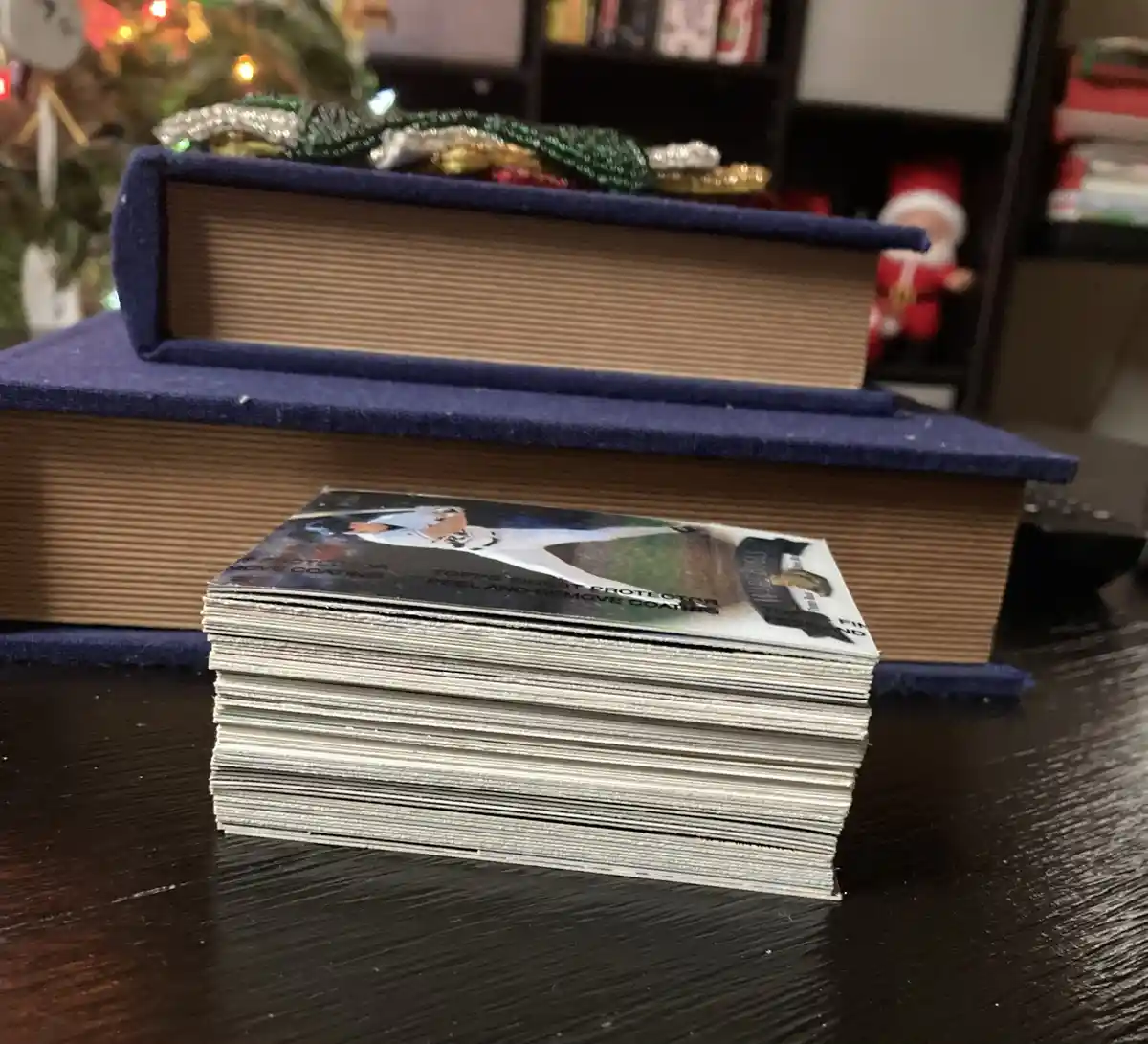 A thick stack of baseball trading cards sits on a dark wooden table, with two large books stacked behind it. The top card…