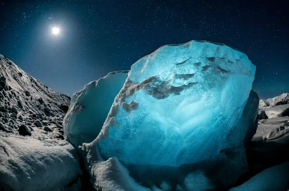 A large, translucent blue ice block glows under a bright moon and starry night sky, surrounded by snow-covered terrain. The…
