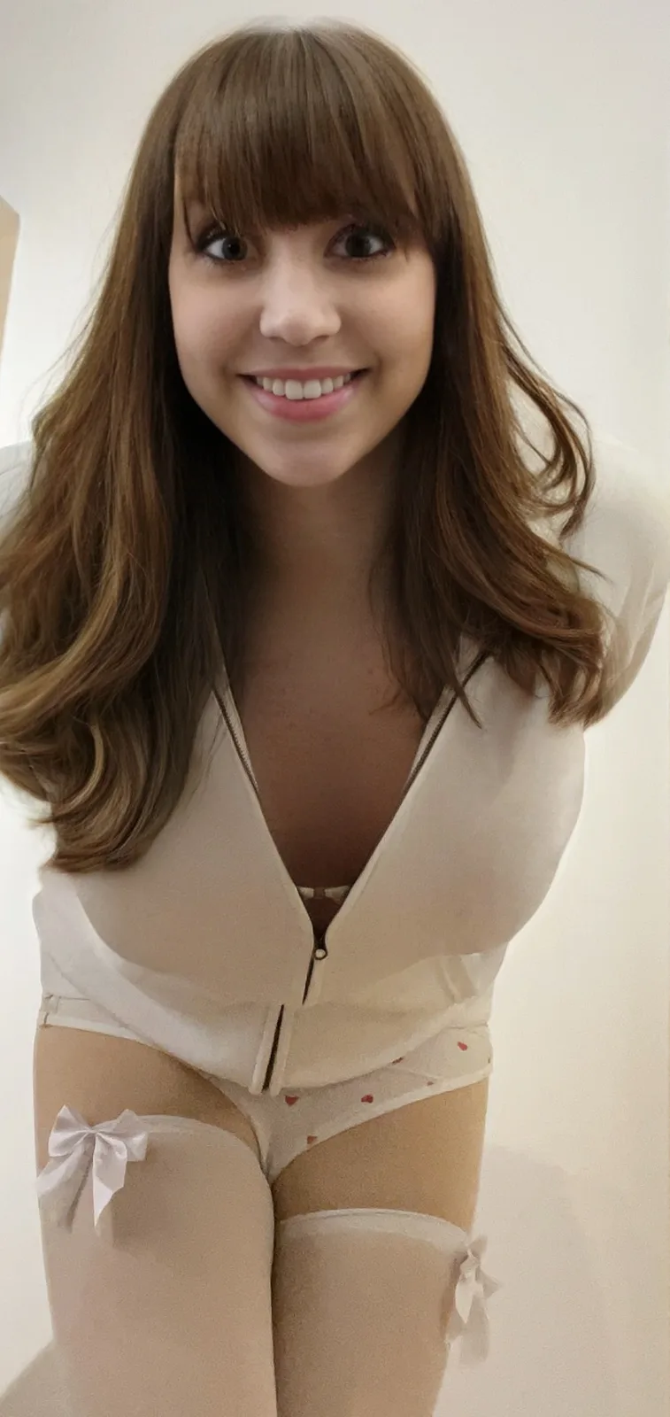 A young woman with brown hair and bangs smiles at the camera while wearing a sheer white bodysuit with a zipper down the…