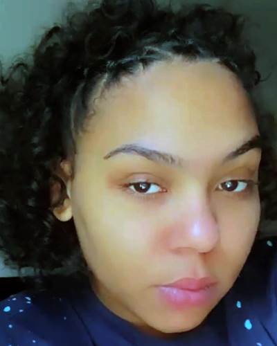 A close-up selfie of a woman with dark curly hair and a neutral expression, wearing a dark blue shirt with light blue polka…