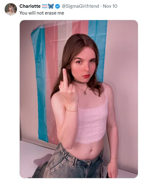 A young woman with brown hair and red lipstick stands in front of a trans pride flag, pointing her middle finger directly at…