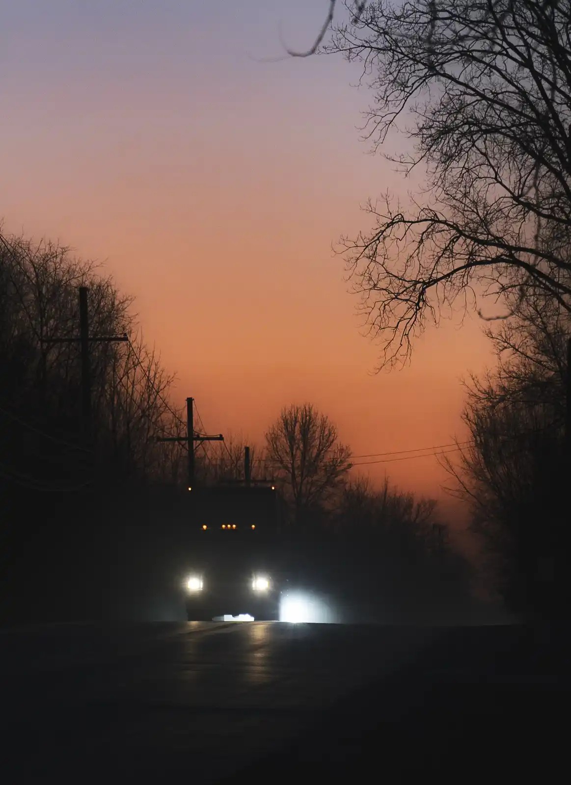 A dark, atmospheric photo of a large truck approaching on a rural road at dusk, its headlights piercing the gloom against a…