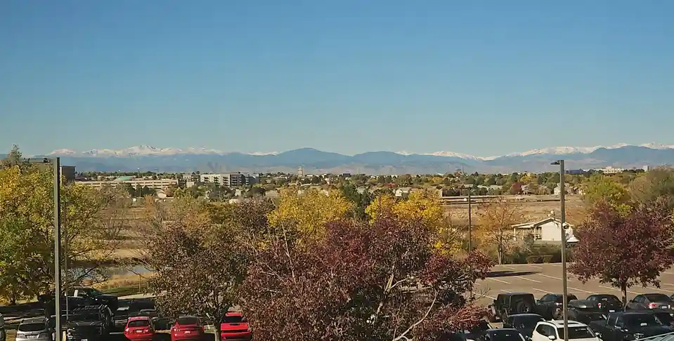 A scenic photo shows a cityscape with a parking lot in the foreground, autumn-colored trees, and a distant mountain range…