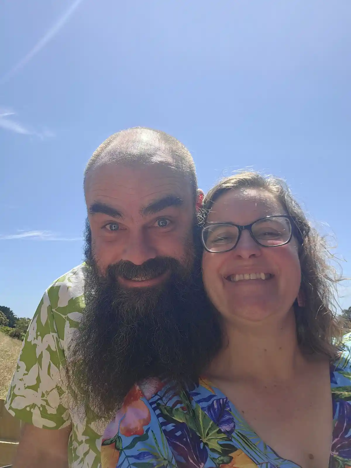 A selfie of a man with a long beard and a woman with glasses, both smiling under a bright blue sky. The man is wearing a…