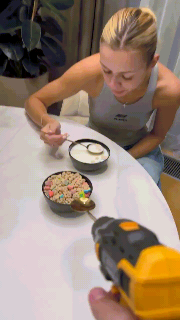 A woman sits at a table with two bowls of cereal, one with colorful cereal pieces and one with white yogurt. A hand holding…
